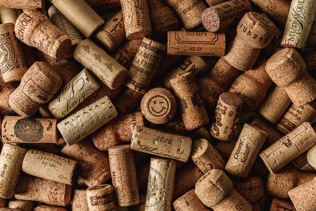 A close-up image of a pile of wine corks. The corks display a variety of labels, logos, and text, indicating different wineries and vintages. Among the corks, one stands out with a smiling and winking face drawn on it. The collection includes both regular wine corks and champagne corks, creating a textured and detailed composition that reflects the enjoyment of wine