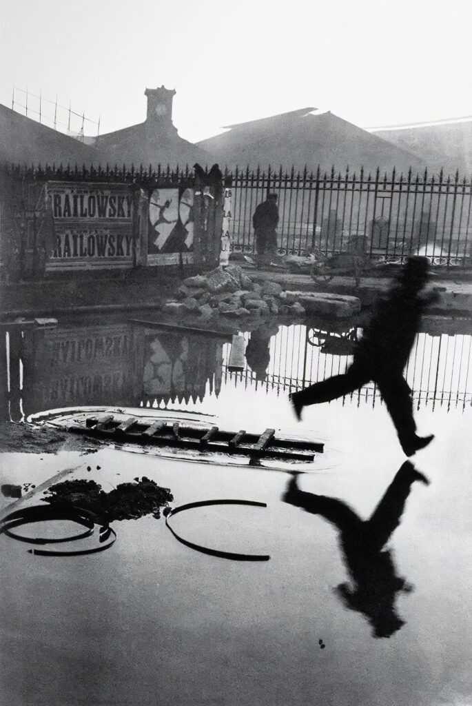 This image is a black-and-white photograph depicting a man in mid-air, captured while leaping over a large puddle. His shadow and reflection are visible in the water below. In the background, there is a metal fence, and behind it, a building with posters on it. One of the posters reads “RAILOWSKY.” The scene has a somewhat industrial or urban feel, with construction materials like a wheelbarrow and piles of rubble scattered around. The photograph has a strong contrast between light and dark areas, creating a dramatic and striking visual effect.