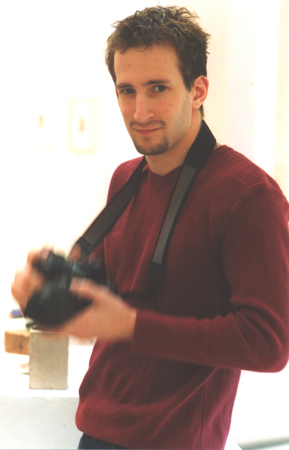 A young man with short, slightly tousled hair is standing indoors, holding a camera. He is wearing a maroon sweater and has a camera strap around his neck. The background is softly lit and slightly blurred, suggesting an indoor setting with some art pieces or decorations visible in the distance. The man has a slight smile on his face and is looking directly at the camera.
